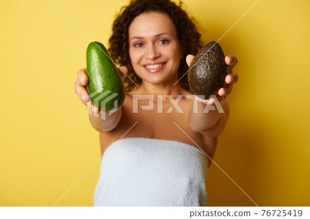 Soft focus on ripe ready-to-eat avocado fruit in hands of blurry half-naked curly smiling woman wrapped in towel. Soft focus on ripe ready-to-eat avocado fruit in hands of blurry half-naked curly smiling woman wrapped in towel. 76725419