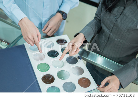 the hands of a shopkeeper and a customer by finger pointing to a sample of lenses when selecting the type of lens 76731108