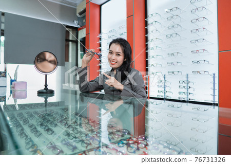 a beautiful woman is posing holding glasses against the background of a glasses display case 76731326