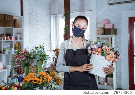 young female florist wearing apron and face mask holding bucket flower smiling looking at camera. working in flower shop young female florist wearing apron and face mask holding bucket flower smiling looking at camera. working in flower shop 76734800