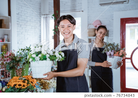 happy young man florist wearing apron holding bucket flower smiling looking at camera. working in flower shop 76734843
