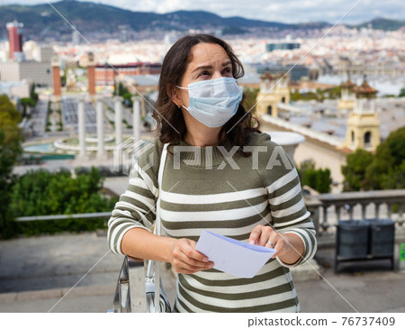 Girl traveler in protective mask with a guidebook in her hands on the street of european city 76737409
