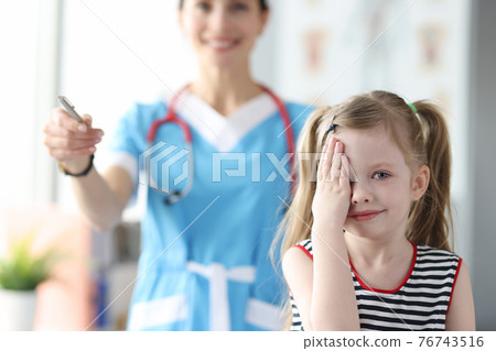 Little girl covering her eyes with her hand at ophthalmologist appointment Little girl covering her eyes with her hand at ophthalmologist appointment 76743516