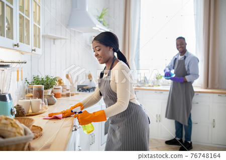 Cheerful black woman cleaning kitchen table, her boyfriend washing dishes on background, blank space 76748164