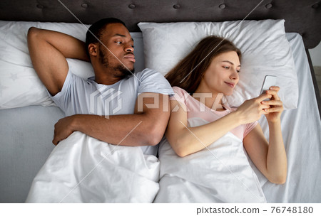 Irritated black guy feeling jealous of his girlfriend stuck in smartphone in bed, top view. Phubbing concept Irritated black guy feeling jealous of his girlfriend stuck in smartphone in bed, top view. Phubbing concept 76748180