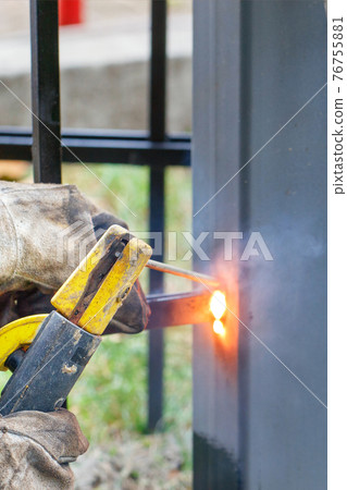 The hands of a welder in rough leather gloves with an electrode holder are welding a metal fence. 76755881