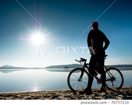 Young man staying on the shore of the lake with bicycle at sunset Young man staying on the shore of the lake with bicycle at sunset 76757210