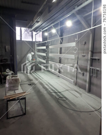 A male painter in a white disposable protective suit paints a metal structure with a spray gun at a paint factory 76758198