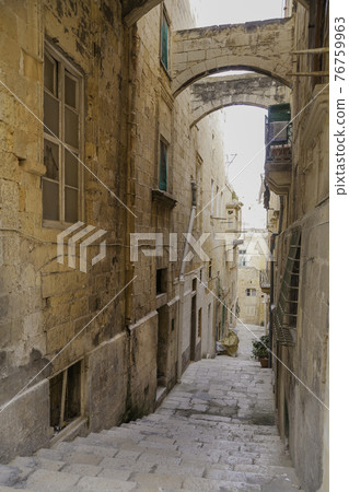 Ancient narrow street in Valletta, Malta. Limestone buildings with old fashioned lanterns and balconies. Staircase with many steps as pavement. Ancient narrow street in Valletta, Malta. Limestone buildings with old fashioned lanterns and balconies. Staircase with many steps as pavement. 76759963