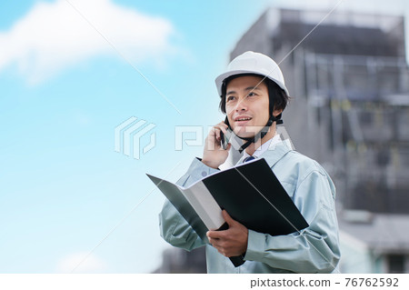 A man in work clothes talking on a smartphone in front of a construction site 76762592