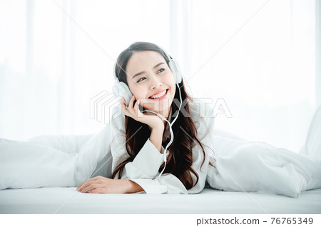 Beautiful young asian woman in headphones listening to music and lying down on bed 76765349