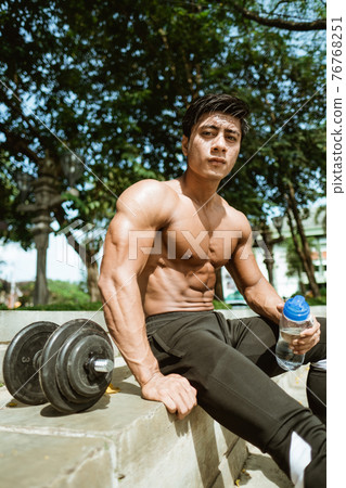 The shirtless muscular man sits holding a drinking bottle by the dumbbells after a hand muscle workout The shirtless muscular man sits holding a drinking bottle by the dumbbells after a hand muscle workout 76768251