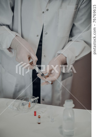 Female hands in white medical gloves drawing medicine from ampoule into a syringe, next to upright ampoule with medicine on white acrylic table. 76769615