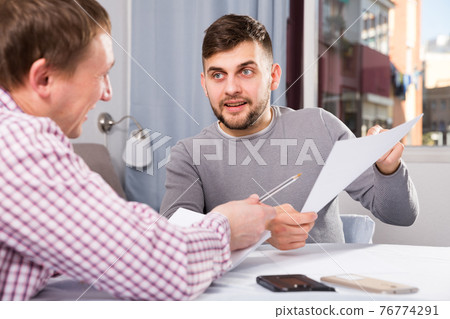 Cheerful man reading documents with friend at home table Cheerful man reading documents with friend at home table 76774291