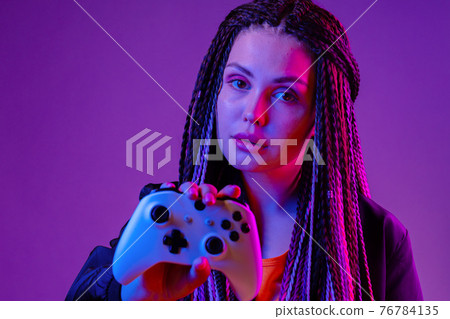 Young woman with long braids holding console joystick against dark purple background Young woman with long braids holding console joystick against dark purple background 76784135