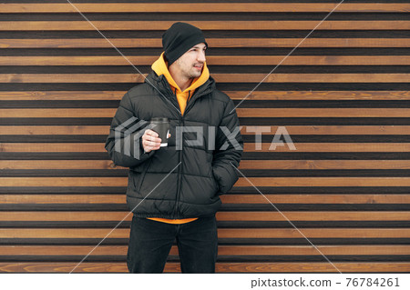 Young man in winter clothes standing with coffee to go in a street against wooden wall Young man in winter clothes standing with coffee to go in a street against wooden wall 76784261