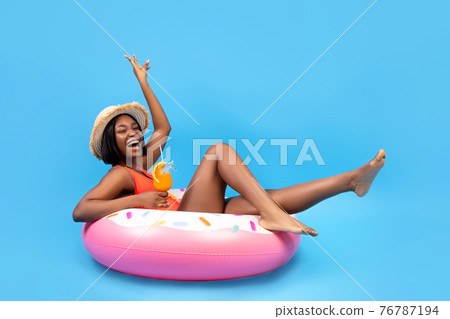 Positive black woman sitting on inflatable ring, holding tropical cocktail on blue background, full length portrait 76787194