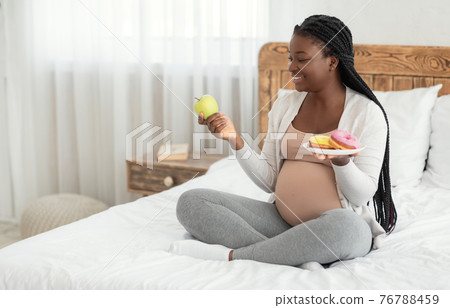 Prenatal Nutrition. Black Pregnant Woman Choosing Between Apple And Donuts At Home 76788459