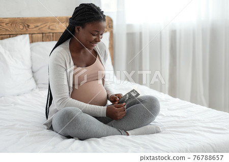 Happy black pregnant woman with ultrasound picture of baby sitting on bed 76788657