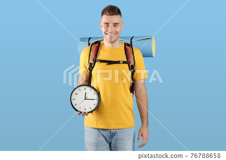Time to travel concept. Handsome millennial guy with backpack holding clock over blue studio background 76788658
