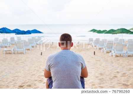 Young guy sits with his back on a sandy beach Young guy sits with his back on a sandy beach 76790260