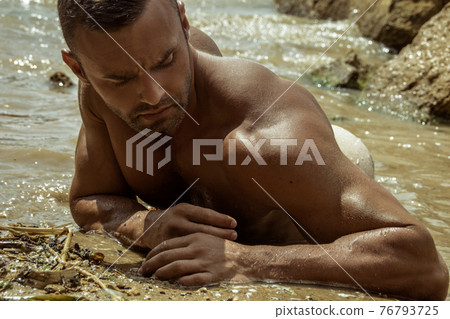 Strong face muscle male model on the nature background with sand stones and water on the beach on the sunset Strong face muscle male model on the nature background with sand stones and water on the beach on the sunset 76793725
