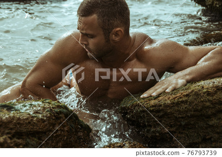 Strong face muscle male model on the nature background with sand stones and water on the beach on the sunset Strong face muscle male model on the nature background with sand stones and water on the beach on the sunset 76793739