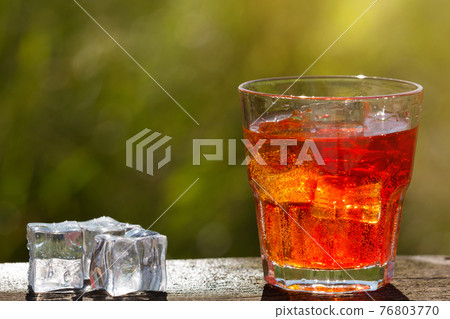 a glass with an exotic red drink and ice cubes on the boards, against a background of greenery 76803770