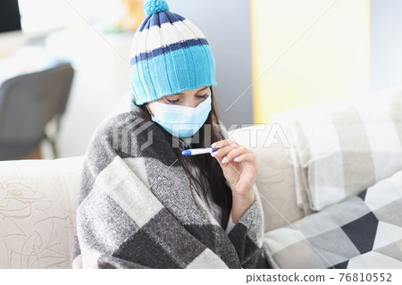 Woman in cap and protective medical mask looking at thermometer Woman in cap and protective medical mask looking at thermometer 76810552