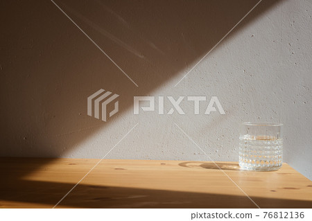 A glass with clean clear water and sharp shadows stands on a wood table on gray wall background 76812136