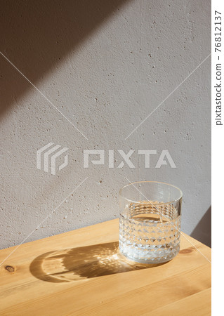 A glass with clean clear water and sharp shadows stands on a wood table on gray wall background A glass with clean clear water and sharp shadows stands on a wood table on gray wall background 76812137