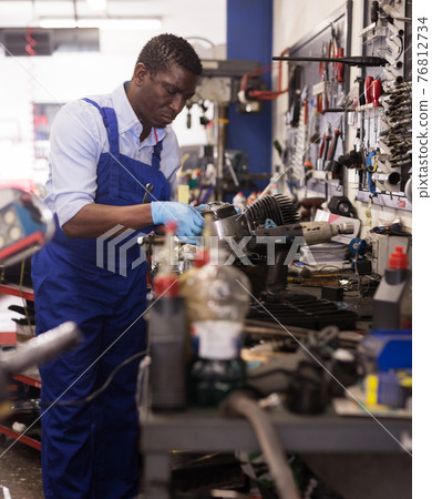 Mechanic in overalls repairing a motorcycle engine in a garage Mechanic in overalls repairing a motorcycle engine in a garage 76812734