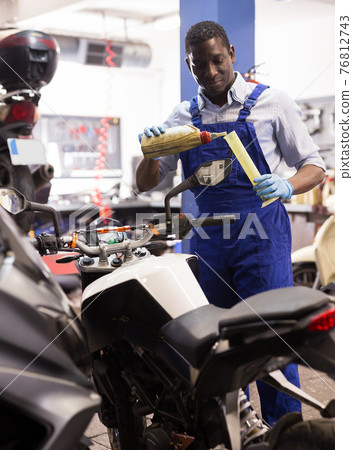 Engineer in overalls pours oil for motorcycle engine in garage 76812743