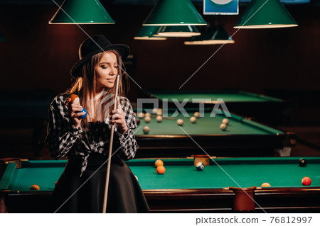 A girl in a hat in a billiard club with a cue in her hands.billiards Game 76812997