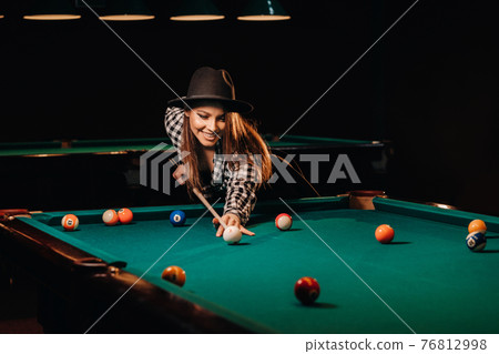 A girl in a hat in a billiard club with a cue in her hands hits a ball.Playing billiards 76812998