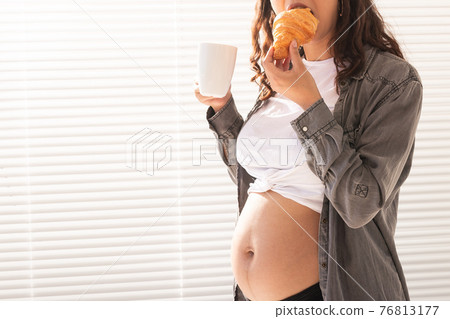 Healthy beautiful pregnant woman drinking tea and eating croissant during lunch. Concept of high-calorie nutrition while waiting for birth of baby. Copy space. 76813177