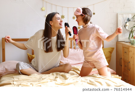 Enjoying weekend at home. Cute happy girl little daughter with hairbrush as mic singing with mom while sitting together on bed 76814944