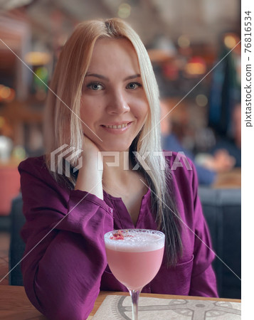 A blonde girl in a lilac sweater sits in a cozy restaurant on a sofa and she is relaxed and have a pink cocktail on her table.. 76816534