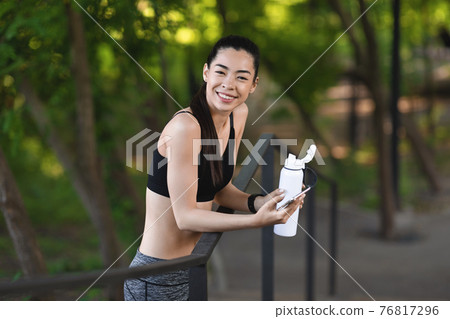 Sporty asian woman taking break after running outdoors, holding smartphone and water Sporty asian woman taking break after running outdoors, holding smartphone and water 76817296