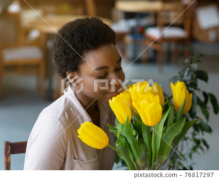 Portrait of beautiful black lady smelling bouquet of yellow tulips at coffee shop 76817297
