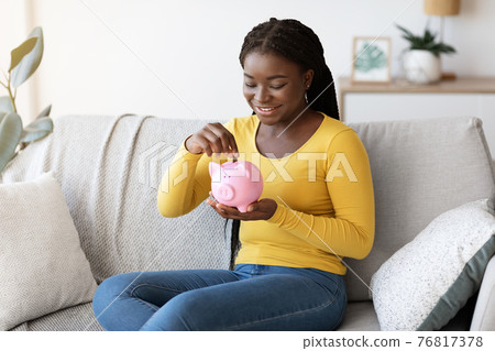 Black woman putting coin in piggy bank, sitting on couch at home 76817378