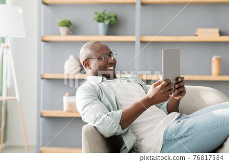 African american man reading e-book on digital tablet or browsing internet, enjoying weekend and resting on armchair African american man reading e-book on digital tablet or browsing internet, enjoying weekend and resting on armchair 76817584