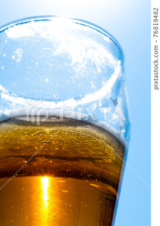 Light beer in a glass goblet on a background of blue sky. Beer foam on the walls of a glass in the sunlight. 76819482