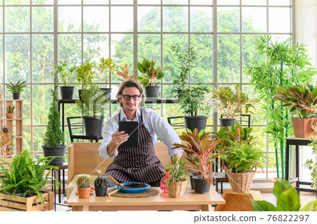 Happy senior man gardening wearing glasses and denim apron with digital tablet sitting in home gardening for looking who buys the tree. Small business concept 76822297