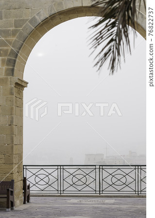 Valletta through morning fog and architectural arch and palm tree leaf. View from Upper Barrakka Garden. Misty weather in capital of Malta. 76822737