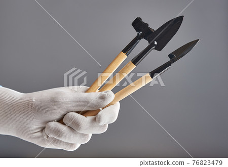 Hand in new pure white glove holding gardening tools isolated on gray background. Hand in new pure white glove holding gardening tools isolated on gray background. 76823479