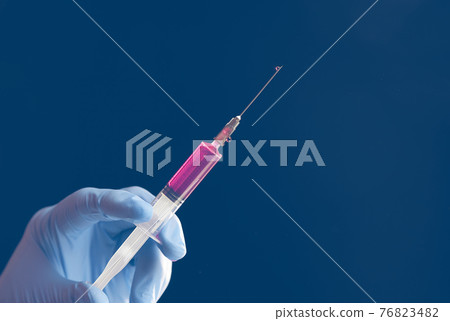 Doctor's blue-gloved hand holding a syringe with red liquid isolated on blue background. Doctor's blue-gloved hand holding a syringe with red liquid isolated on blue background. 76823482