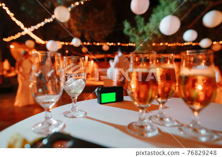 Black digital camera stands on a table covered with a white tablecloth against a background of glasses with drinks Black digital camera stands on a table covered with a white tablecloth against a background of glasses with drinks 76829248