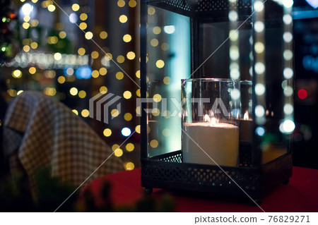 iron lantern with glass inside a candle is burning with a flame of glowing warm light in the night backyard with bokeh from festive garlands on the window in diffocus close-up, nobody. 76829271