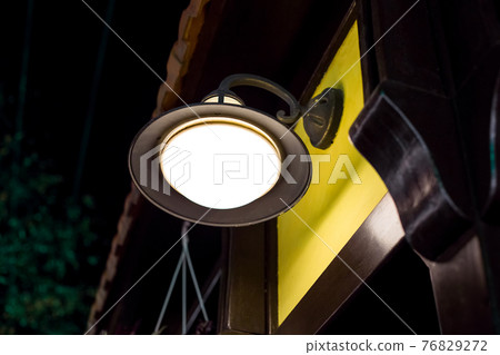 wrought iron wall lamp of round shape and shade with visor on exterior of a building with a yellow facade and wooden architectural elements pilasters and cornice. 76829272